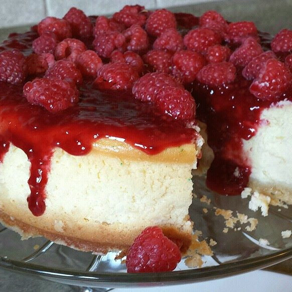 A cheesecake topped with raspberry compote and fresh raspberries, served on a glass cake stand.