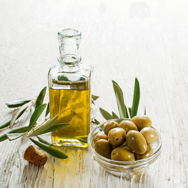 A glass bottle of olive oil beside a bowl of fresh olives, surrounded by olive leaves on a light-colored surface.
