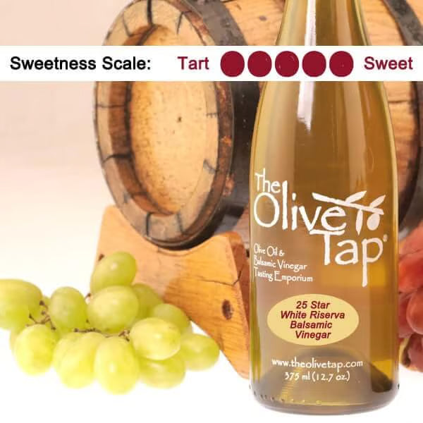 Bottle 25 Star White Riserva Balsamic Vinegar from The Olive Tap with grapes in front of a wooden barrel and a sweetness scale indicating a five out of five.