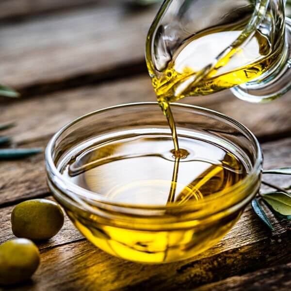 Olive oil being poured from a glass cruet into a clear glass bowl, with olives and olive leaves on a wooden surface.