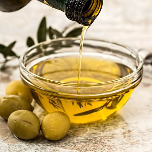 Olive oil being poured into a small glass bowl beside fresh herbs and olives.