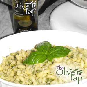 A bowl of pasta tossed with pesto sauce and topped with fresh basil leaves, served in a white dish with an olive oil bottle from The Olive Tap in the background.