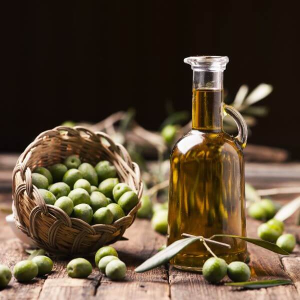 A glass pitcher of olive oil next to a basket of olives and leaves on a wooden surface sprinkled with more olives against a black background.