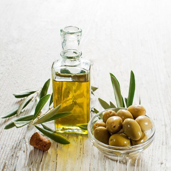 A glass bottle of olive oil with a cork stopper, surrounded by olive branches and a bowl of green olives on a wooden surface.