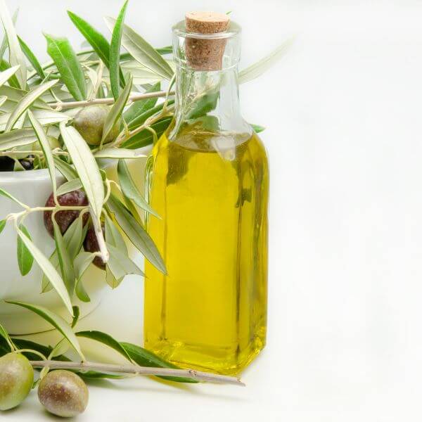 A glass bottle of olive oil with a cork stopper, displayed beside olive branches and green olives against a white background.