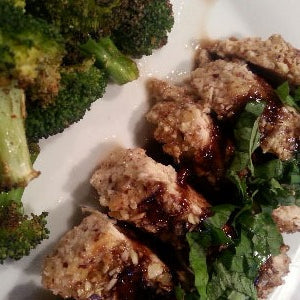 A sliced almond crust chicken breast topped with fresh basil and balsamic glaze, served with roasted broccoli on a white plate.