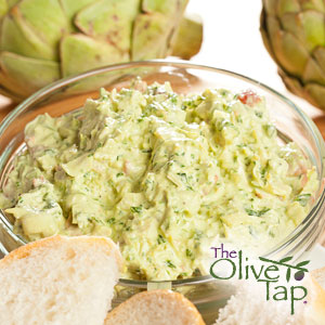 A glass bowl of artichoke parmesan dip with sliced French bread with artichokes in the background.