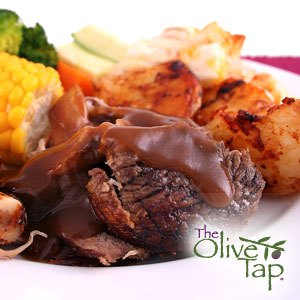 Baked lamb topped with mole sauce, served alongside potatoes, corn on the cob, and broccoli on a white plate.