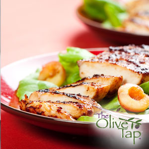 A sliced chicken breast with a balsamic glaze on leafy greens with a halved peach on each on each side on a red and white plate on a red table.