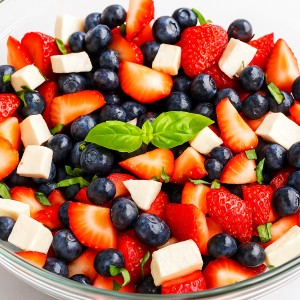 A salad of blueberries, cubed mozzarella cheese, strawberries, and fresh basil pieces garnished with a basil leaf and served in a glass bowl.