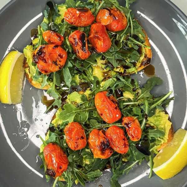 Two slices of avocado toast topped with arugula and blistered tomatoes and drizzled with a balsamic vinegar on a gray plate with lemon wedges.