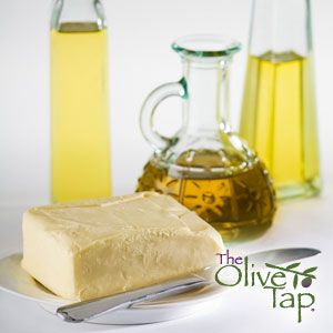 A block of butter on a dish beside a knife with three glass bottles of olive oil in the background on a white surface and white background.