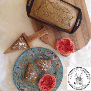Grapefruit and cardamom olive oil cake in a pan next to a halved grapefruit and cake slices dusted with powdered sugar served on a decorative plate.