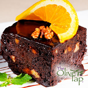 Chocolate brownie with walnut chunks, topped with a glossy chocolate glaze, orange slice, and walnut pieces, served on a white plate with a mint garnish.