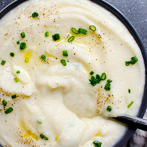 Creamy cauliflower mash topped with spices, olive oil, and chives in a black bowl with a spoon.