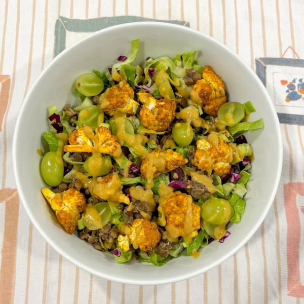 Curried cauliflower on a bed of spring mix, lentils, and halved green grapes, drizzled with a creamy dressing in a white bowl.