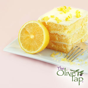 A square slice of lemon layered cake with a lemon cream cheese frosting topped with fresh lemon zest on a white plate with an upside down for and a lemon sliced in half.