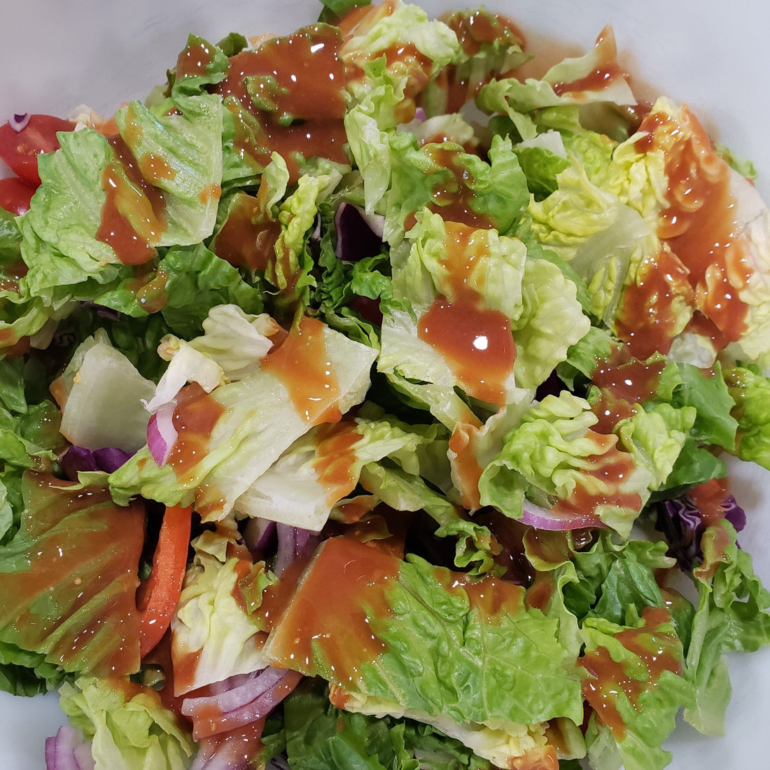 A mixed green salad with romaine lettuce, red onion, tomatoes, purple cabbage, and dressing served in a white bowl.