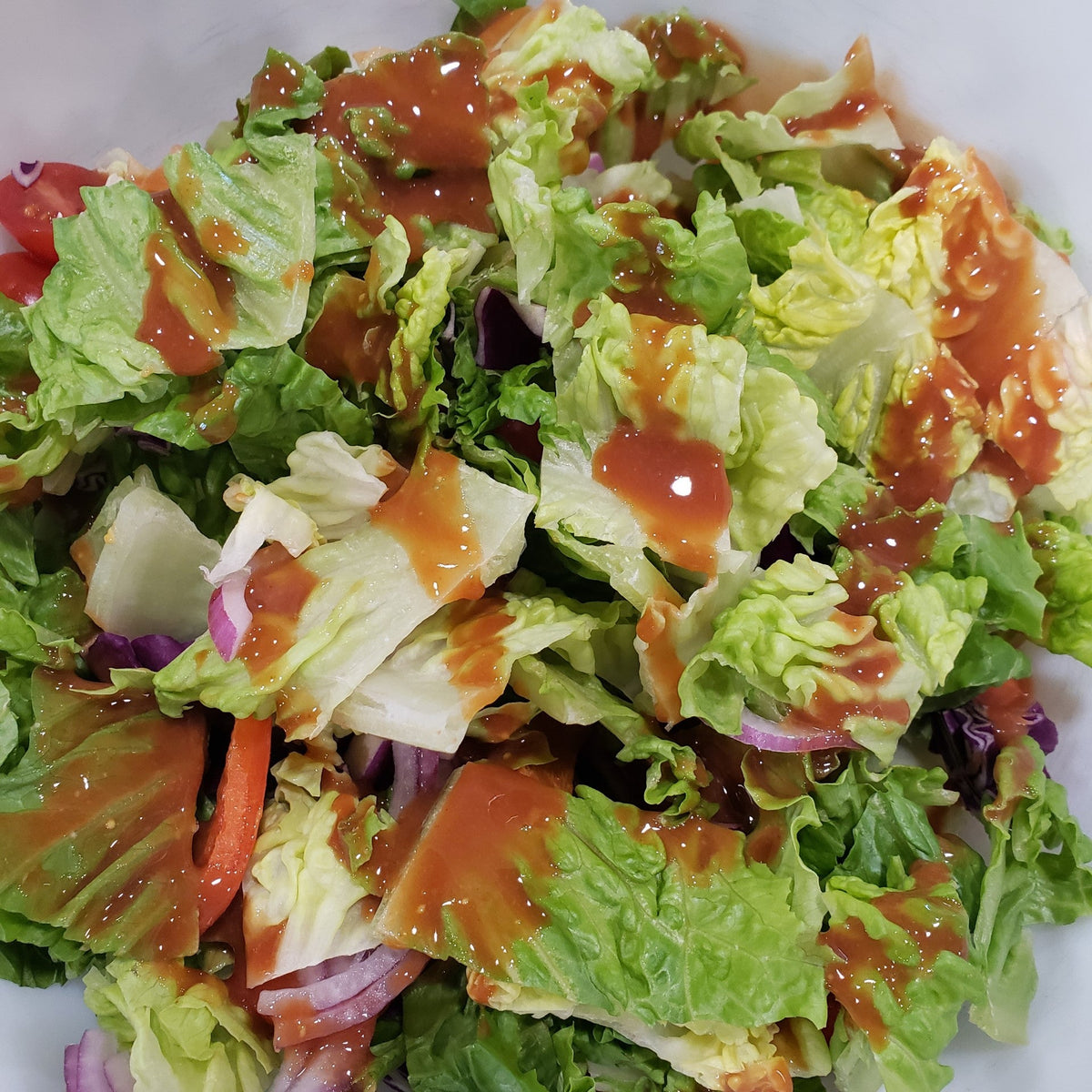 A mixed green salad with romaine lettuce, red onion, tomatoes, purple cabbage, and dressing served in a white bowl.