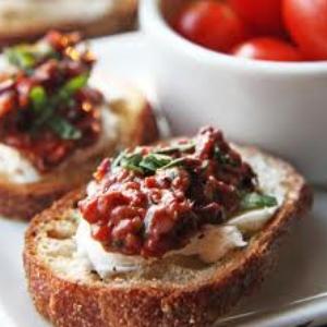 Crostini topped with a creamy cheese spread and sun-dried tomato mixture, garnished with herbs and served on a white plate.
