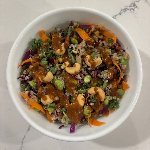 Quinoa salad with carrots, cashews, red cabbage, scallions, edamame and drizzled with peanut sauce in a white bowl.