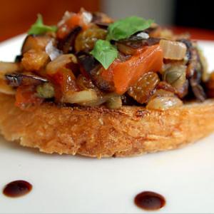 A slice of toasted bread topped with a mixture of eggplant, tomatoes, golden raisins, onions, and fresh basil, served on a white plate with dots of a dark sauce.