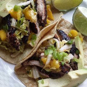 Tacos filled with grilled chicken, cabbage mango slaw, and avocado slices, garnished with green onion, and served with lime wedges on a white plate. 