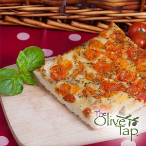 A large piece of focaccia bread topped with cherry tomatoes and herbs on a cutting board with fresh basil and cherry tomatoes in a basket.