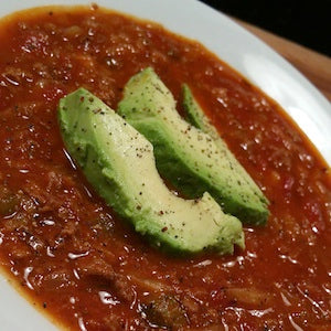 A bowl of red broth soup with ground beef and tomato chunks, topped with sliced avocado and black pepper.