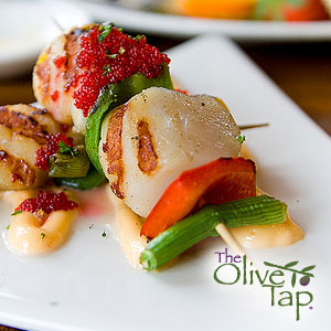 A grilled skewer with scallops, green onion, and red pepper topped with red fish eggs on a squiggle of creamy sauce and served on a white plate.