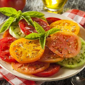 Green, red, and yellow heirloom tomato slices stacked on a white plate, sprinkled with seasoning and garnished with fresh basil and a red and white checkered napkin underneath the plate.