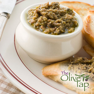 A green olive tapenade in a white bowl on a white plate with sliced bread, one piece topped with the tapenade.