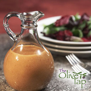 A glass pitcher filled halfway with vinaigrette with a mixed greens salad and forks in the background on a gray surface and maroon background.