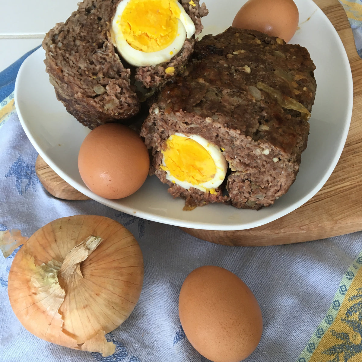Two meatloaves stuffed with hard-boiled eggs on a white plate, surrounded by eggs and an onion.