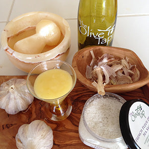 A glass of Catalan Allioli surrounded by a bottle of olive oil, garlic bulbs, sea salt, and mortar and pestle on a wooden board.