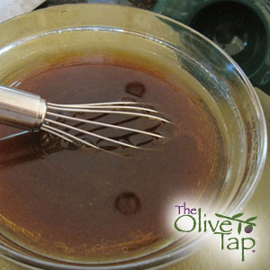 A vinaigrette in a glass mixing bowl with a whisk inside and The Olive Tap logo on the righthand corner.