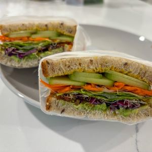 Fresh vegetable sandwich on whole grain bread, layered with avocado, cucumber, shredded carrots, leafy greens, and purple cabbage, cut in half and served on a white plate.