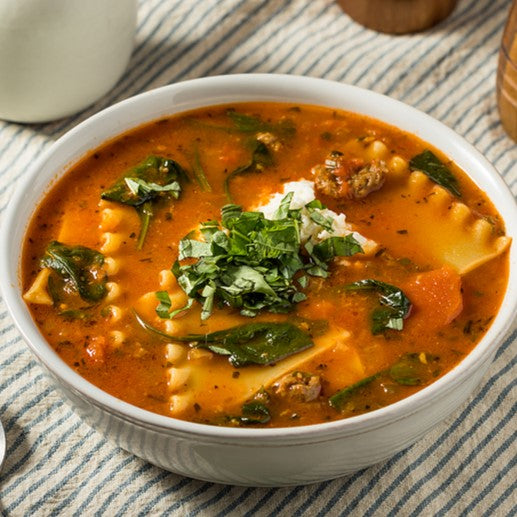 Tomato-based soup filled with lasagna noodles, sausage, topped with fresh herbs and ricotta cheese in a white bowl.
