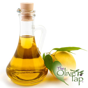 Lemon vinaigrette in a glass dressing bottle next to a fresh lemon with its leaves on a white background and The Olive Tap logo on the bottom righthand corner.