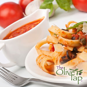 Pasta tossed in a red tomato sauce and sliced olives served on a white plate next to a fork and a sauce serving dish of red tomato sauce with fresh garlic, tomatoes, and basil in the background on a white surface.