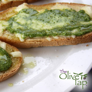 Toasted garlic bread topped with melted cheese and a generous layer of vibrant green pesto, served on a white plate.