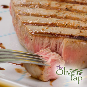 Close-up of a grilled tuna steak with grill marks and a tender pink center, being flaked with a fork on a white plate.