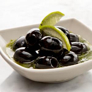 Marinated black olives in an herbal olive oil, served in a white bowl and garnished with lime slices.