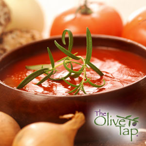 Tomato soup garnished with fresh rosemary in a wooden bowl surrounded by shallots and fresh tomatoes.