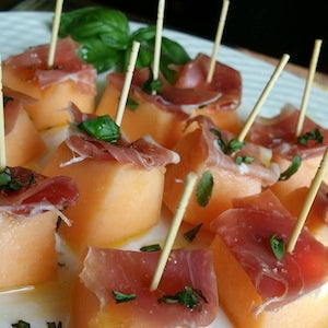 Cantalope cubes topped with prosciutto and fresh basil held together with toothpicks, served on a white plate.