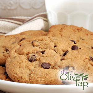 Freshly baked chocolate chip cookies arranged on a white plate, with a glass of milk and a folded kitchen towel in the background.
