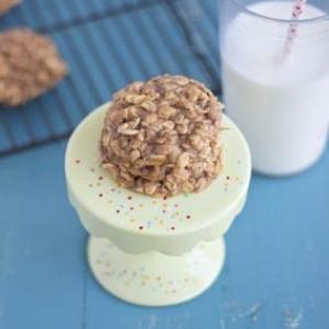 An oatmeal cookie served on a small cake stand, with a glass of milk in the background.