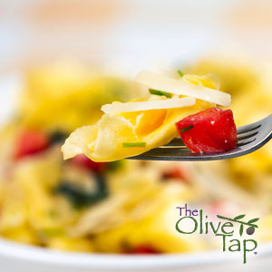 A close-up of a noodle and red pepper with parmesan cheese on a fork with a bowl of the pasta blurred in the background.