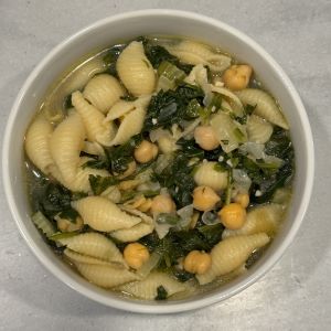 Bowl of vegetable pasta soup with shell-shaped pasta, chickpeas, leafy greens, onions, and herbs in a light broth and served in a white bowl on a light-colored surface.