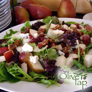 Mixed greens salad topped with crumbled prosciutto, sliced pears, red grapes, crumbled goat cheese, and walnuts, served on a white platter.